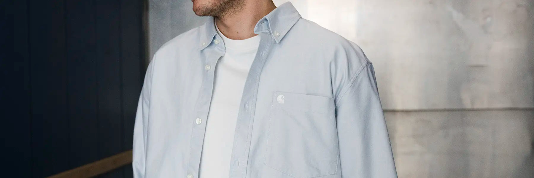 A person in a light blue button-up shirt from Carhartt WIP over a white T-shirt stands against a textured gray and dark blue wall, exuding a casual, relaxed style.