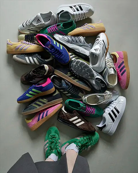 A collection of various Adidas sneakers arranged artistically, with a pair of green shoes worn by a person in the foreground.